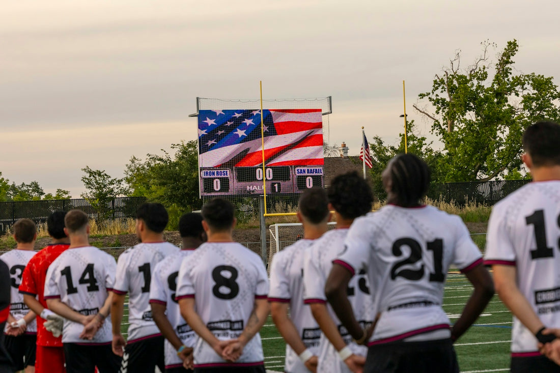 Soccer players stand facing a scoreboard with american flag.