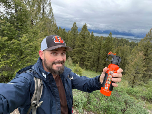 Man holds bear spray in a mountain forest landscape.