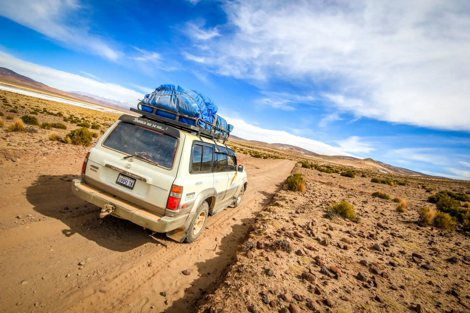 A white SUV with a roof rack loaded with blue tarps, equipped for off-road travel.
